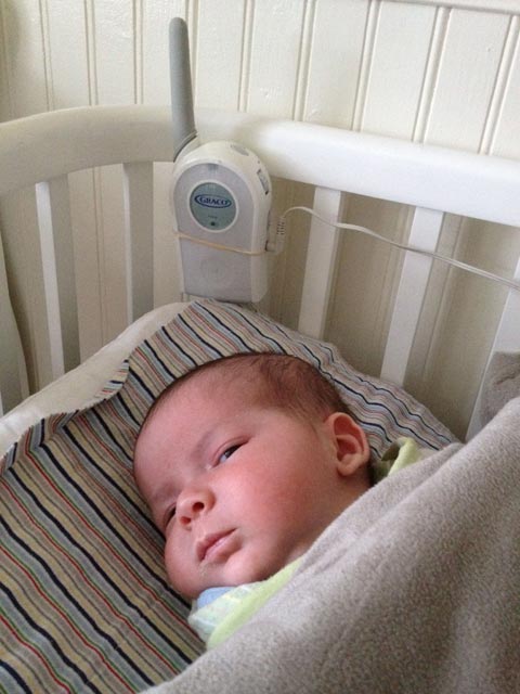 Baby boy Max looks askance from his white Argington Bam Bassinet, with a Graco baby monitor tied to the side with a rubber band