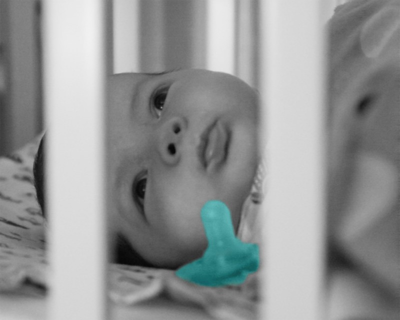 baby boy Max seen through the bars of his BAM bassinet, with a pacifier by his face. The image is grayscale with a colorful blue-green pacifier.
