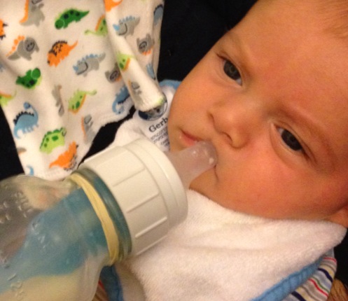 sam, wearing a white bib, has clearly had enough milk and won't open his mouth for more. The nipple is pressed against his lips and he has a stubborn expression.