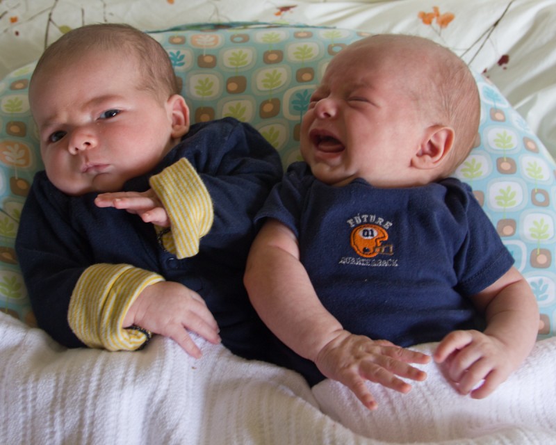 twin baby boys max and sam sit together on a Boppy pillow, both wearing navy blue. sam cries toward max and max recoils