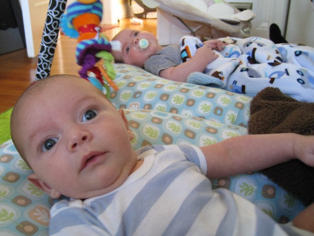 baby boys sam and max sit in Boppy Newborn Loungers under a play structure with dangling toys. Sam in the foreground appears to be talking.