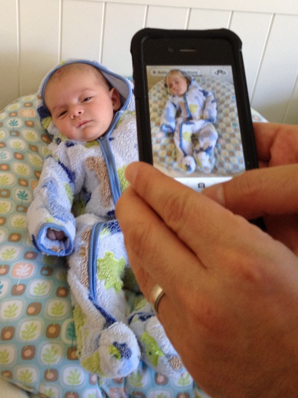 baby boy sam sits wearing a zip-up suit while his father takes a photo of him with an iPhone. The photo, taken with an iPhone, is of a photo being taken with an iPhone