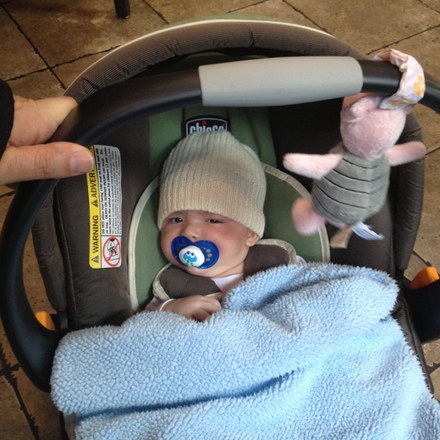 baby boy max sits in a Chicco car seat, with a pacifier in his mouth and his knit cap rolled out surfer-style. Piglet hangs from above.