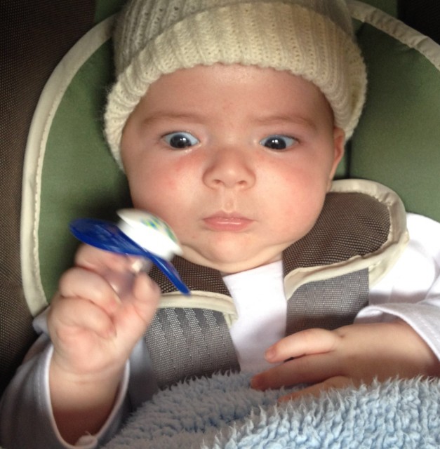 baby boy max sitting in a Chicco car seat and wearing a knit cap, holds onto a pacifier that he has removed from his mouth and he stares at it with great focus